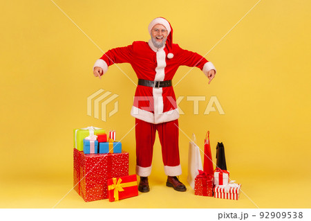 Full length portrait of smiling elderly man with gray beard wearing santa claus costume pointing at many present boxes for Christmas holidays. Indoor studio shot isolated on yellow background. 92909538