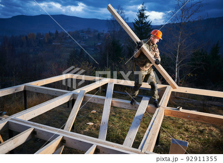 Man worker building wooden frame house on pile foundation. Carpenter carrying wooden board for timber framing. Carpentry concept. Man worker building wooden frame house on pile foundation. Carpenter carrying wooden board for timber framing. Carpentry concept. 92911355