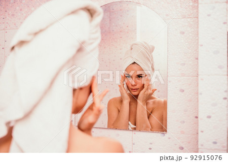 Young beautiful woman using face cream moisturizing lotion after bath. Pretty attractive girl in a towel on her head stands in front of a mirror in a home bathroom. Daily hygiene and skin care Young beautiful woman using face cream moisturizing lotion after bath. Pretty attractive girl in a towel on her head stands in front of a mirror in a home bathroom. Daily hygiene and skin care 92915076