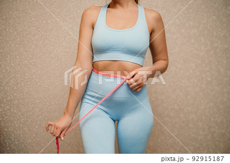 Cropped view of slim woman measuring waist with tape measure at home, close up. Unrecognizable european woman checking the result of diet for weight loss or liposuction indoors 92915187