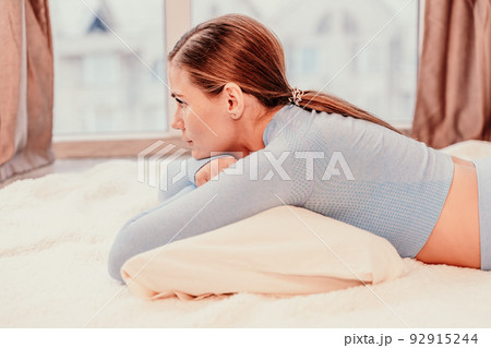 Side view portrait of relaxed woman with long hair lying on carpet at home. She is dressed in a blue tracksuit, holding a phone in her hands. 92915244