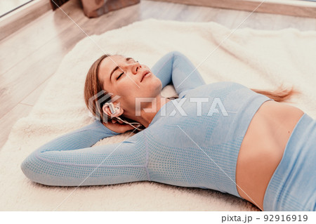 Top view portrait of relaxed woman listening to music with headphones lying on carpet at home. She is dressed in a blue tracksuit. 92916919