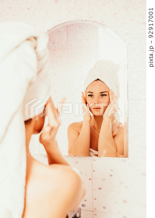 Young beautiful woman using face cream moisturizing lotion after bath. Pretty attractive girl in a towel on her head stands in front of a mirror in a home bathroom. Daily hygiene and skin care 92917001