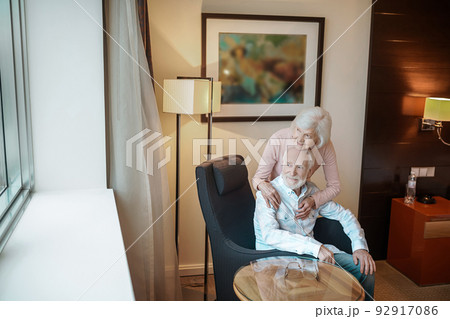 Short-haired senior woman hugging her husband while he sitting in the chair 92917086