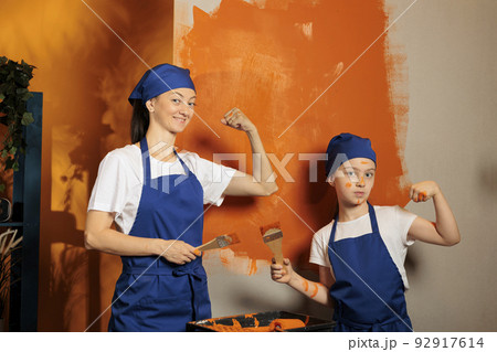 Portrait of mom and kid flexing arm muscles while painting room walls with orange color paint and brush. Woman with little child using renovating tools, working together on house decor. 92917614
