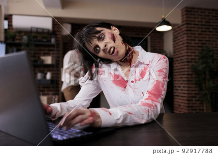 Aggressive devil corpse looking at laptop, sitting at office desk and being dangerous. Brain eating woman monster with bloody gory wounds being dramatic and eerie with mouth open. 92917788