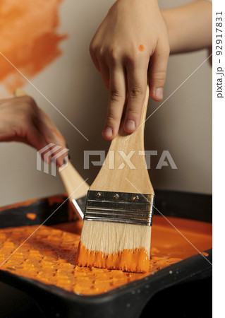 Small family using paintbrush and color to paint room walls, holding brush with bristles in liquid acrylic can. People painting apartment with paintwork tools, surface design renewal. Close up. Small family using paintbrush and color to paint room walls, holding brush with bristles in liquid acrylic can. People painting apartment with paintwork tools, surface design renewal. Close up. 92917831