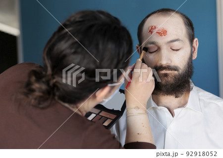 Artist using makeup effects to create zombie costume and scary dramatic corpse look. Man looking deceased and infected after having bloody scars and monster face, creepy design. 92918052
