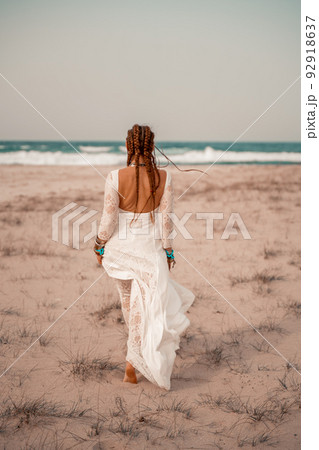Model in boho style in a white long dress and silver jewelry on the beach. Her hair is braided, and there are many bracelets on her arms. 92918637