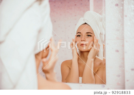 Young beautiful woman using face cream moisturizing lotion after bath. Pretty attractive girl in a towel on her head stands in front of a mirror in a home bathroom. Daily hygiene and skin care 92918833