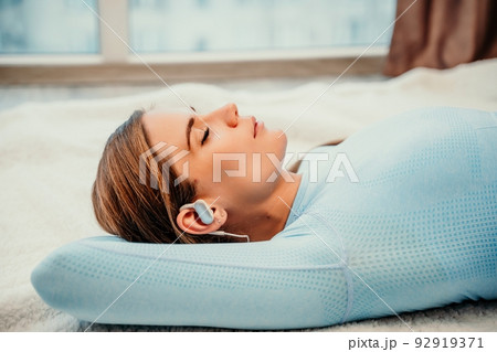 Side view portrait of relaxed woman listening to music with headphones lying on carpet at home. She is dressed in a blue tracksuit. 92919371