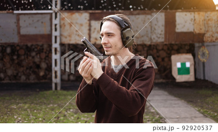 A young man shoots a gun, aiming at the target. A man wearing protective headphones. A wall and a roof with bullet holes. outdoor shooting range 92920537