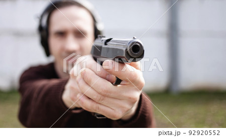 A young man is aiming for a gun. A man wearing protective headphones. outdoor tyre shooting range. hobby. firearm against the white wall A young man is aiming for a gun. A man wearing protective headphones. outdoor tyre shooting range. hobby. firearm against the white wall 92920552