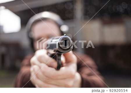 A young man shoots a gun, aiming at the target. A man wearing protective headphones. A wall and a roof with bullet holes. outdoor shooting range A young man shoots a gun, aiming at the target. A man wearing protective headphones. A wall and a roof with bullet holes. outdoor shooting range 92920554