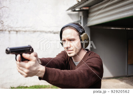 A young man is aiming for a gun. A man wearing protective headphones. outdoor tyre shooting range. hobby. firearm against the white wall 92920562