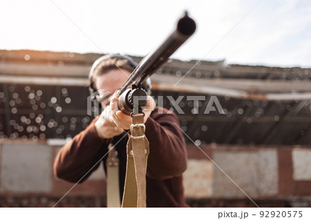 A young man in protective glasses and headphones. A pump-action shotgun. Tyre's outdoors. A wall and a roof with bullet holes. Sport A young man in protective glasses and headphones. A pump-action shotgun. Tyre's outdoors. A wall and a roof with bullet holes. Sport 92920575