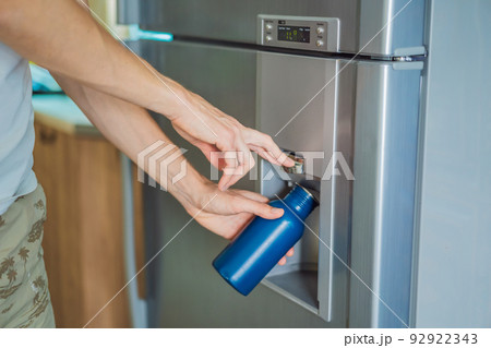 Male hand is pouring cold water and ice cubes in a metal bottle from dispenser of home fridge 92922343