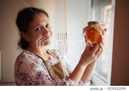 Hispanic pretty woman, pleasant housewife, stands by the window at home kitchen, smiles at the camera, presenting homemade peach jam, confiture, marmalade, canned according traditional family recipe Hispanic pretty woman, pleasant housewife, stands by the window at home kitchen, smiles at the camera, presenting homemade peach jam, confiture, marmalade, canned according traditional family recipe 92930269