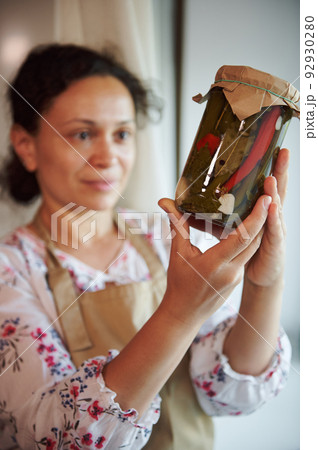 Focus on a glass jar of homemade pickled red and green chili peppers, marinated according a traditional family recipe in the hands of a multiethnic cute housewife, standing by a window at home kitchen Focus on a glass jar of homemade pickled red and green chili peppers, marinated according a traditional family recipe in the hands of a multiethnic cute housewife, standing by a window at home kitchen 92930280