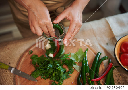 Overhead view of a housewife in a beige kitchen apron, filling sterilized glass jar with spicy chili peppers and fresh chopped fragrant ingredients when pickling seasonal vegetables in the kitchen Overhead view of a housewife in a beige kitchen apron, filling sterilized glass jar with spicy chili peppers and fresh chopped fragrant ingredients when pickling seasonal vegetables in the kitchen 92930402