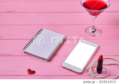 Smartphone with blank screen and open notepad for copy space. Womens accessories on pink wooden desk. 92931015