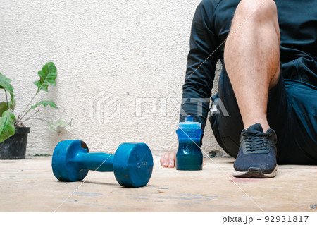 man sitting on the floor next to blue dumbbell and bottle with water, after exercising. exercise at home, white background. fitness concept. health concept, space for advertising text. 92931817