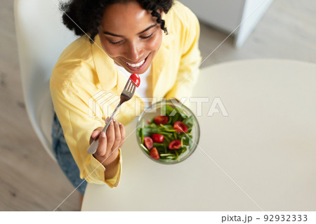 Fit african american lady eating salad with organic vegetables in light kitchen interior, top view, free space Fit african american lady eating salad with organic vegetables in light kitchen interior, top view, free space 92932333