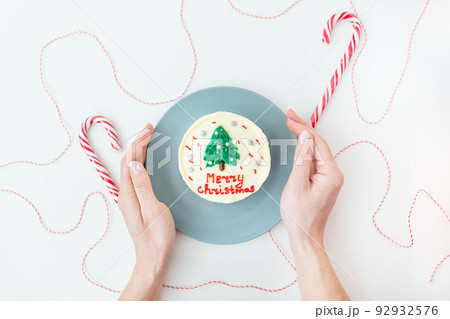 A girl holds a plate with a Christmas cake with the inscription Merry Christmas, decorated with icing, on a white background with lollipops and a red thread for gifts. 92932576