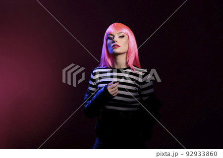 Caucasian attractive woman advertising punk fashion trend in dark shadow light, standing over pink background. Beautiful stylish person wearing leather jacket and pink hair wig in studio. 92933860