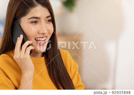 At home in the living room.Asian woman talks on the phone on the sofa in a calm attitude in the morning.Calling pals and having hanppy and good time. At home in the living room.Asian woman talks on the phone on the sofa in a calm attitude in the morning.Calling pals and having hanppy and good time. 92935898