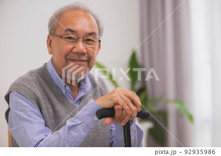 Asian Elderly man holding walking stick for physiotherapy.Sitting on sofa after walking in morning. Asian Elderly man holding walking stick for physiotherapy.Sitting on sofa after walking in morning. 92935986