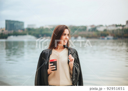 Young woman with take away coffee in hand in casual clothes near river water Young woman with take away coffee in hand in casual clothes near river water 92951651