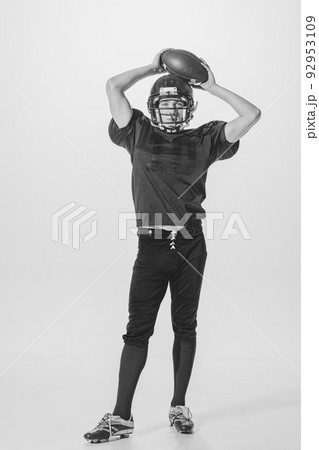 Portrait of young man, american football player posing in helmet and uniform with ball. Black and white photography Portrait of young man, american football player posing in helmet and uniform with ball. Black and white photography 92953109