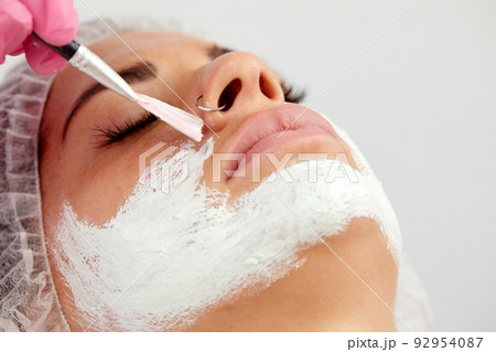 Satisfied young Caucasian beautiful woman uses services of professional beautician in beauty salon or spa. Unrecognizable beautician applied white mask to half of client's face with brush. Top view. 92954087