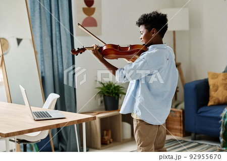 Rear view of African boy standing in his room and practicing to play violin during online lesson on laptop Rear view of African boy standing in his room and practicing to play violin during online lesson on laptop 92955670