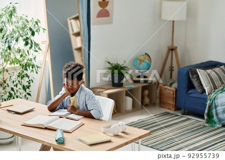 African little boy sitting at desk in his room and preparing his homework reading a book African little boy sitting at desk in his room and preparing his homework reading a book 92955739