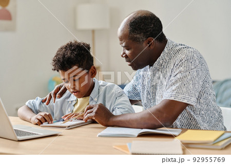 African teacher explaining new material to schoolboy pointing at notebook while they sitting at desk in the room 92955769