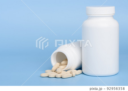 Two white medicine bottles, one closed and second with pills spilled on blue background Two white medicine bottles, one closed and second with pills spilled on blue background 92956358