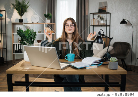 Woman freelancer in eyeglasses sitting at table with closed eyes and relieving stress by meditation 92958994