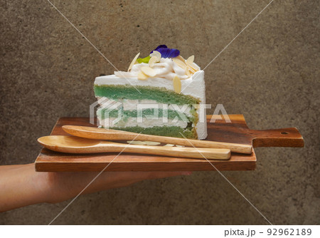hold brown wooden chopping board, butterfly pea cake with three layer of cake on top by coconut and a butterfly pea flower, sprinkle with almond slices and two light brown wooden spoons, on cement hold brown wooden chopping board, butterfly pea cake with three layer of cake on top by coconut and a butterfly pea flower, sprinkle with almond slices and two light brown wooden spoons, on cement 92962189