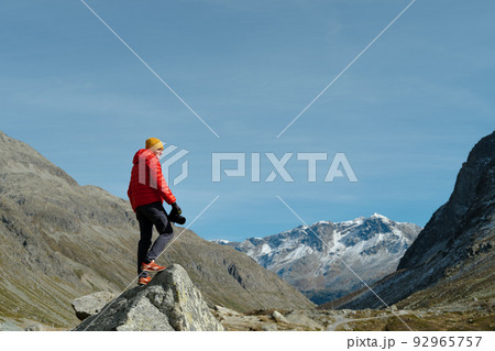 Back view of a tourist man in red jacket and camera standing with a view on Julier Pass, Switzerland Back view of a tourist man in red jacket and camera standing with a view on Julier Pass, Switzerland 92965757