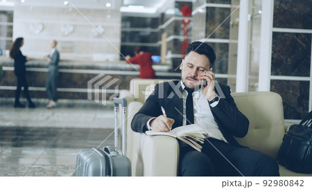 Pan shot of confident businessman talking mobile phone and writing notes in notepad while sitting on armchair in luxury hotel with luggage near him. Travel, business and people concept 92980842