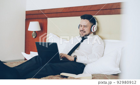 Happy cheerful businessman in headphones working at laptop computer and listening music smiling while lying in bed at hotel room. Travel, business and people concept 92980912