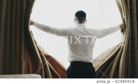 Businessman open curtains in hotel room at the morning and looking into window Businessman open curtains in hotel room at the morning and looking into window 92980918