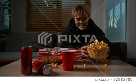 Happy man watching TV and holding bowl of popcorn while sitting on couch at home. Leisure activity, relaxation, hobby concept Happy man watching TV and holding bowl of popcorn while sitting on couch at home. Leisure activity, relaxation, hobby concept 92985700