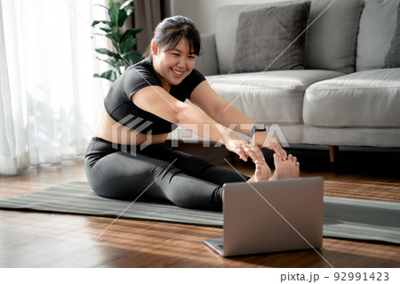 Asian chubby woman sitting on the floor in living room practice online yoga lesson with the computer. female having meditate training class on the laptop.. Asian chubby woman sitting on the floor in living room practice online yoga lesson with the computer. female having meditate training class on the laptop.. 92991423