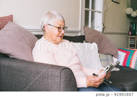 Old senior woman, 80s adult, grandmother, making crossword puzzles in her living room sitting on the sofa. Retirement hobby for training memory. Alzheimer's preventive practice. Selective focus 92991607