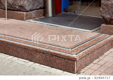 entrance with granite threshold with non-slip rubber to shop from pedestrian sidewalk from plink on footmat through glass door, rough stone facade of building close-up architecture, nobody. 92994717