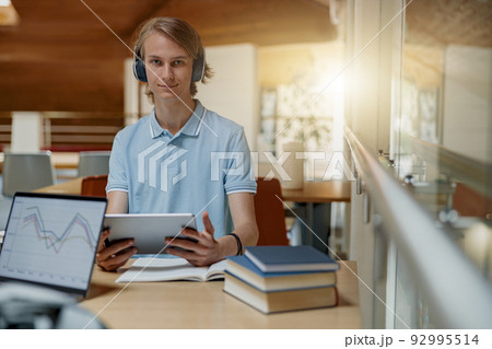 Smiling Male student sits in headphones in library and studies using a digital tablet Smiling Male student sits in headphones in library and studies using a digital tablet 92995514