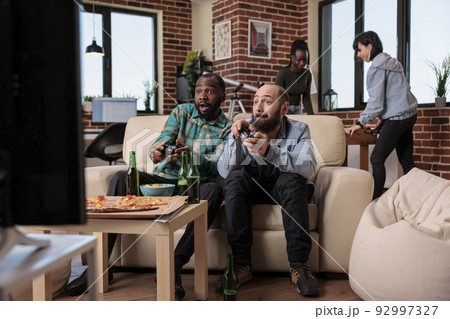 Diverse friends having fun with video gaming competition on tv console, playing online games at party gathering with beer bottles and pizza. Enjoying hangout with people, entertainment. 92997327